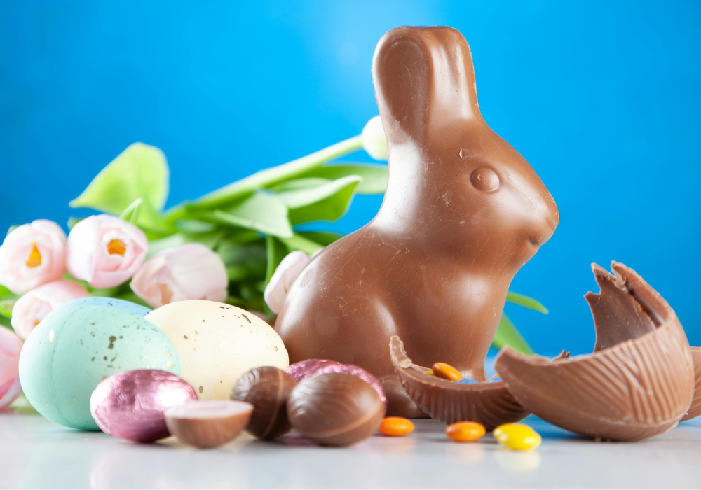 A Chocolate Easter Rabbit and Chocolate Easter Eggs and sweets on a table with a bouquet of light pink tulips, and a blue wall behind